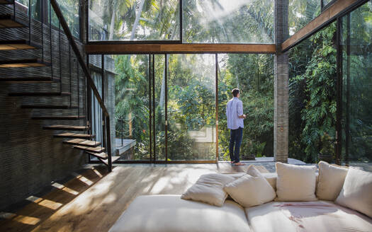 Thoughtful young man standing by glass door at home - IKF00754