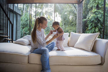 Mother with baby daughter standing on sofa at home - IKF00757