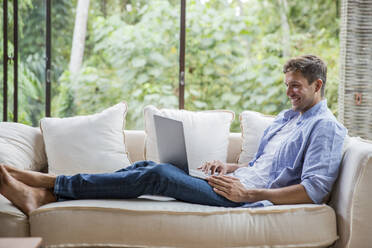 Happy freelancer using laptop on sofa at home - IKF00778