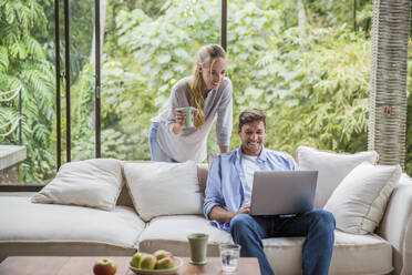 Happy freelancer using laptop with woman holding coffee cup at home - IKF00791