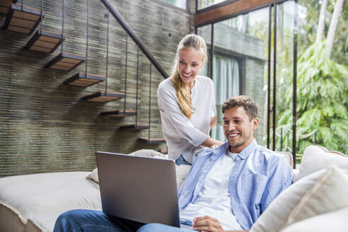 Happy freelancer using laptop with woman on sofa at home - IKF00793