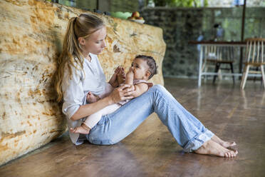 Mother with daughter sitting on floor in kitchen at home - IKF00799