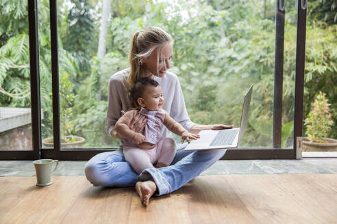 Happy freelancer using laptop with baby girl sitting at home - IKF00805