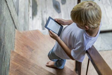 Blond boy using e-reader and standing on staircase at home - IKF00809