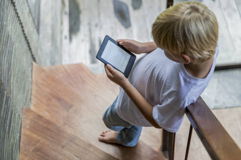 Blonder Junge, der einen E-Reader benutzt und zu Hause auf einer Treppe steht - IKF00809