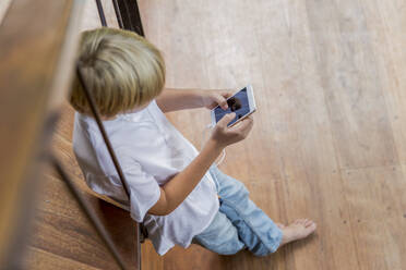 Blond boy using smart phone and leaning on railing at home - IKF00810