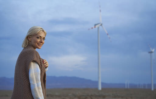 Blonde Frau im Pullover steht in der Nähe von Windrädern in der Wüste - AZF00535