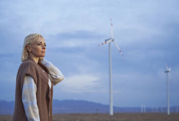 Blond woman with hand in hair standing near wind turbines in desert - AZF00536