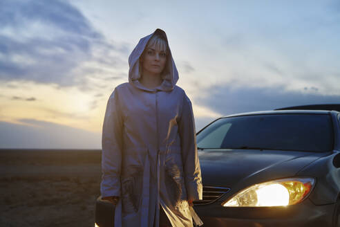 Blonde Frau im Regenmantel steht neben dem beleuchteten Scheinwerfer eines Autos - AZF00538