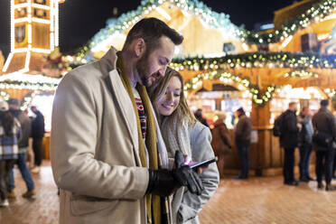 Junge Frau mit Mann, der auf dem Weihnachtsmarkt ein Smartphone benutzt - WPEF07358
