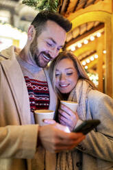 Glückliches Paar mit Smartphone und Kaffeetasse auf dem Weihnachtsmarkt - WPEF07369