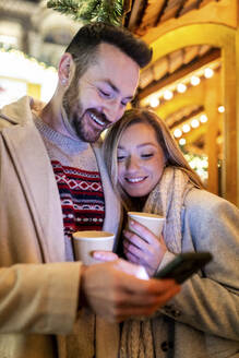 Happy couple using smart phone and holding coffee cup at Christmas market - WPEF07369