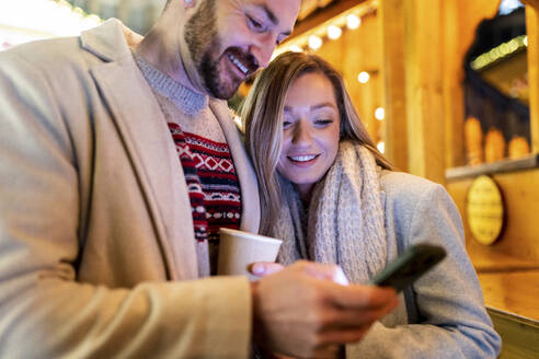 Smiling woman and man using mobile phone at Christmas market - WPEF07370