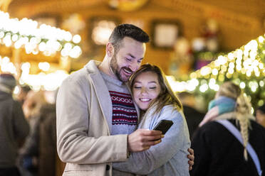 Ein glücklicher Mann und eine Frau benutzen ein Mobiltelefon auf dem Weihnachtsmarkt - WPEF07389