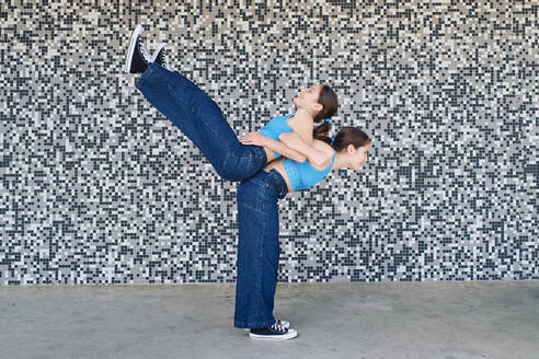 Happy girl carrying sister on back in front of textured wall - ASGF03603