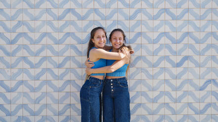 Glückliche Zwillingsschwestern, die sich vor einer Wand umarmen - ASGF03630