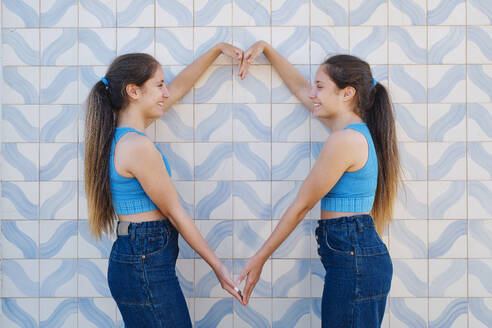 Happy sisters gesturing heart shape in front of wall - ASGF03637