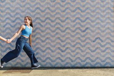 Happy girl holding hand of sister and running in front of wall - ASGF03641