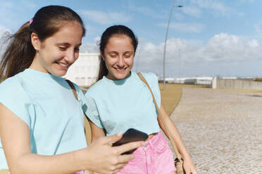 Happy twin sisters using smart phone on sunny day - ASGF03646