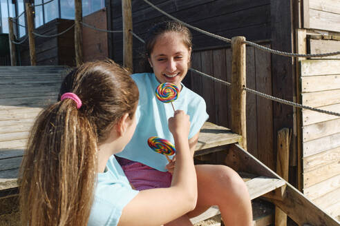 Happy sisters with lollipops sitting on sunny day - ASGF03650