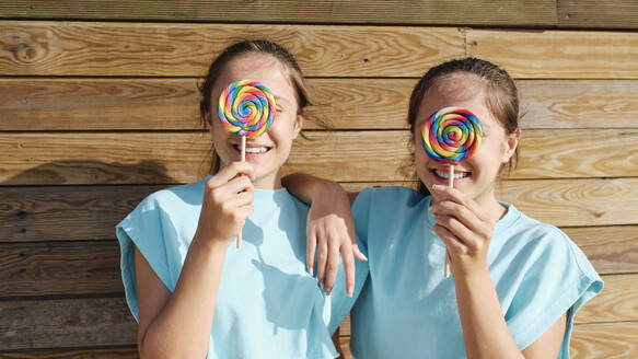 Happy sisters holding lollipop over face in front of wooden wall - ASGF03653