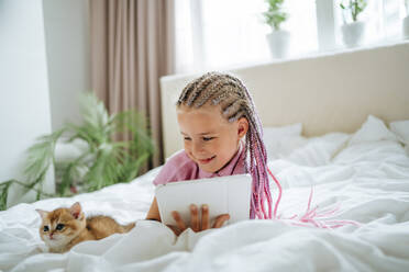 Girl holding tablet PC lying by kitten on bed - MDOF01220