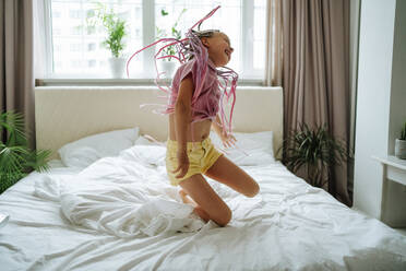 Glückliches Mädchen mit rosa Dreads, das auf dem Bett hüpft - MDOF01224