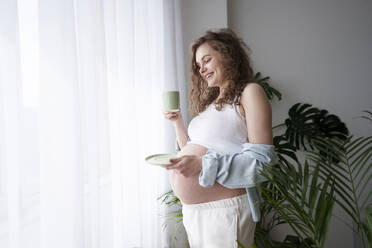 Smiling pregnant woman having coffee near window at home - AAZF00640