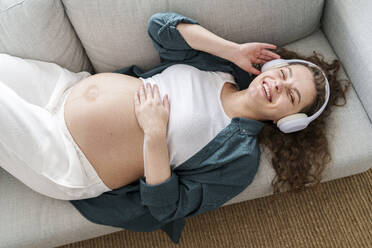 Happy pregnant woman listening to music lying down on sofa at home - AAZF00651