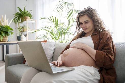 Junge schwangere Frau mit Laptop zu Hause sitzend - AAZF00652