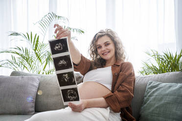 Lächelnde schwangere Frau mit Ultraschallbildern auf dem Sofa zu Hause - AAZF00655