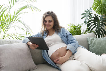Lächelnde schwangere Frau, die einen Tablet-PC benutzt und zu Hause auf dem Sofa sitzt - AAZF00658