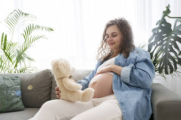 Lächelnde schwangere Frau mit Teddybär auf dem Sofa zu Hause - AAZF00659