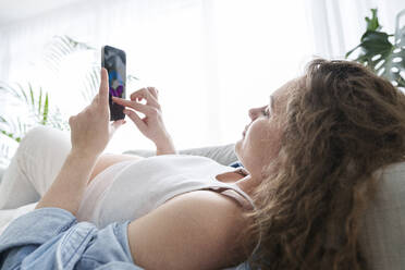 Young pregnant woman using smart phone lying on sofa at home - AAZF00660