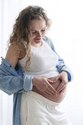 Smiling pregnant woman making heart shape on belly at the window - AAZF00667