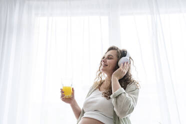 Schwangere Frau hört Musik und trinkt Saft am Fenster - AAZF00674