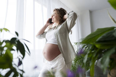 Smiling pregnant woman with hand in hair talking on smart phone at home - AAZF00679