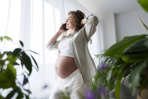 Lächelnde schwangere Frau mit Hand im Haar, die zu Hause mit ihrem Smartphone telefoniert - AAZF00679
