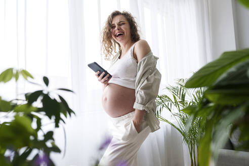 Glückliche schwangere Frau mit Hand in der Tasche hält Smartphone zu Hause - AAZF00680