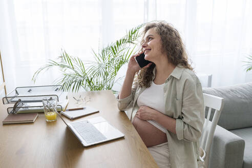 Lächelnde schwangere Freiberuflerin, die im Büro zu Hause mit ihrem Smartphone telefoniert - AAZF00683