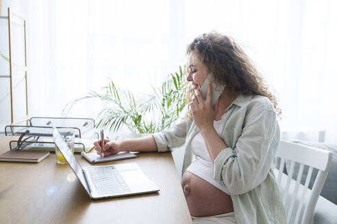 Schwangere Freiberuflerin, die im Büro zu Hause schreibt und mit ihrem Smartphone telefoniert - AAZF00686