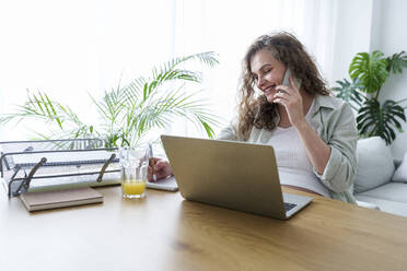 Smiling pregnant businesswoman talking on smart phone at home office - AAZF00687