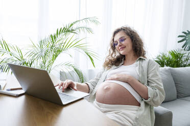 Lächelnde schwangere Freiberuflerin mit Hand auf dem Bauch, die von zu Hause aus arbeitet - AAZF00688