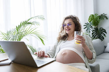 Lächelnde schwangere Freiberuflerin, die einen Laptop benutzt und ein Saftglas im Heimbüro hält - AAZF00689