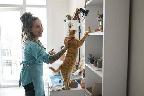 Smiling woman with curious cat looking in shelf - ANNF00254