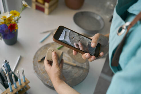 Craftswoman photographing clay cup through smartphone at workbench - ANNF00266