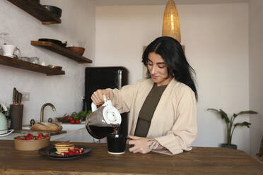 Young woman pouring freshly brewed coffee in cup at kitchen table - SYEF00392