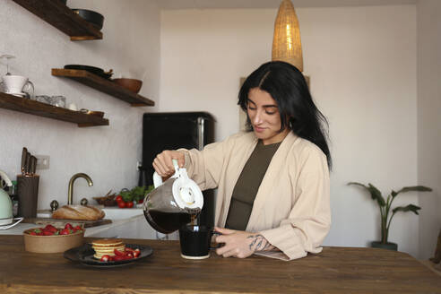 Junge Frau gießt frisch gebrühten Kaffee in eine Tasse am Küchentisch - SYEF00392