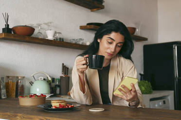 Junge Frau telefoniert und genießt gebrühten Kaffee mit Pfannkuchen am Tisch - SYEF00394
