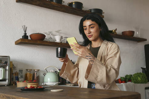 Junge Frau benutzt ein Smartphone und genießt einen Kaffee am Tisch - SYEF00395